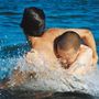 Zwei Männer umarmen sich im Wasser, Wellen spritzen um sie herum, die Szene wirkt intensiv und dynamisch.