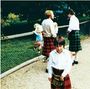 Zwei Männer und ein Junge tragen traditionelle Kilts, stehen vor einem Zaun; im Hintergrund spielen Kinder.