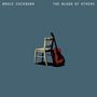 Text: "BRUCE COCKBURN THE BLOOD OF OTHERS". Illustration: Eine Gitarre lehnt an einem Stuhl auf blauem Hintergrund.