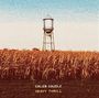 "CALEB CAUDLE HEAVY THRILL" steht vor einem Wasserturm über einem Feld mit hohem, goldenen Gras unter blauem Himmel.