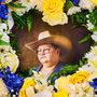Eine Person mit Brille und Cowboyhut, umrahmt von gelben, blauen, weißen Blumen und grünen Blättern.
