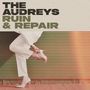 „THE AUDREYS RUIN & REPAIR“; Beine in weißen Hosen, eine Hand hält einen Absatzschuh vor grünem Hintergrund.