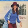 "Carly Simon" und "No Secrets". Eine Frau mit Hut und blauer Tasche steht vor einem dunklen Eisenzaun.