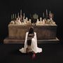 Rückenansicht einer Frau im weißen Kleid mit "ESTRUS" darauf, kniend vor einem Altar mit vielen Kerzen und einer schwarzen Katze.