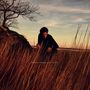 Text: "SIMON MOULLIER CEIBA". Eine Person in einem Feld mit hohem, braunem Gras, ein kahler Baum, Abendhimmel im Hintergrund.