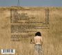Kein Text vorhanden. Eine Person steht neben einem alten Holzhaus in einer weiten, goldenen Feldlandschaft unter bewölktem Himmel.