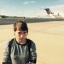 Ich kann den Text nicht erkennen. Eine Frau mit Rucksack steht auf einem Flughafen-Vorfeld, im Hintergrund ein Flugzeug.