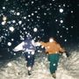 Zwei Personen laufen in winterlicher Landschaft, umgeben von fallendem Schnee, verschwommene Lichter im Vordergrund.
