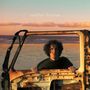Text „mobil home“. Ein Mann steht am Strand hinter einer rostigen Autotür im sandfarbenen Sonnenuntergang.