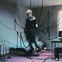 Person in schwarzer Kleidung schwebt mit Kabeln verbunden in einem minimalistisch eingerichteten Zimmer.