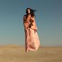 Keine Texte. Eine Frau in einem fließenden, pfirsichfarbenen Kleid steht in einer Wüstenlandschaft.
