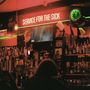 Der Text "SERVICE FOR THE SICK" leuchtet in Neon. Eine Bar mit vielen Flaschen, Zapfhähnen und eine grün beleuchtete Uhr.