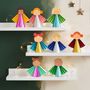 Sechs bunte Engelsfiguren aus Papier stehen auf weißen Regalbrettern, dekoriert mit Lichterketten und Sternen an der Wand.