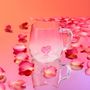 Ein durchsichtiger Becher mit Herzform im Boden, umgeben von rosa Blütenblättern auf einer rosa reflektierenden Oberfläche.
