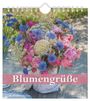 "Blumengrüße 2027." Ein bunter Blumenstrauß mit blauen und rosa Blüten in einer Vase auf einem Holztisch.