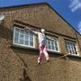 Eine Schaufensterpuppe in rosa Leggings und grauem Oberteil balanciert auf einem Seil vor einem Haus mit brauner Wand und Fenstern.