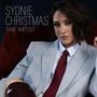 "Sydnie Christmas. The Artist." Eine Person in einem hellblauen Anzug mit roter Krawatte und dunklem Haar schaut ernst.