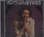 "NATHAN SYKES ULTRAVIOLET". Ein Mann lächelt und faltet die Hände, trägt ein helles Hemd vor dunklem Hintergrund.