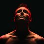 Dunkler Hintergrund, Oberkörper eines Mannes mit nach oben geneigtem Kopf, rot beleuchtet.