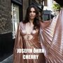 JOSEFIN ÖHRN CHERRY. Frau in schimmerndem, rosafarbenem Kleid, steht auf einer Straße zwischen Gebäuden.