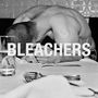 "BLEACHERS" in großen Buchstaben über einem Mann mit gesenktem Kopf an einem Tisch, schwarz-weiß.