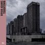 Text: "BRYAN ADAMS TOUGHTOWN". Massive, graue Hochhäuser vor bewölktem Himmel; einsame Person im Vordergrund.