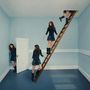 Mehrere Frauen in blauen Kleidern in einem surrealen Raum mit Türen und einer Leiter zur Decke.