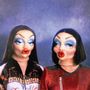 Zwei Figuren mit auffälligem Make-up und puppenartigen Gesichtern, tragen bunte Kleidung.