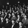 Personen in Anzügen sitzen in mehreren Reihen und tragen alle 3D-Brillen in einem Kino.