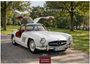 "German Classic Cars" und "2027." Ein weißer Oldtimer mit hochgeklappten Flügeltüren steht auf einer Allee, umgeben von Bäumen.