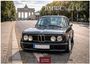 Aufschrift: "BMW Classic Cars". Historisches Auto vor dem Brandenburger Tor, Straße gesäumt von Bäumen.