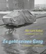 "Es geht seinen Gang" in Blau. "Bertram Kober Fotografien 1977-1990" in Gelb. Auto unter Plane auf grauer Straße.
