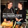 Titel: "Unsere liebsten veganen Alltagsrezepte". Zwei Personen kochen gemeinsam in einer modernen Küche.
