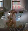 2011-2026, Kate Waters. Eine Frau schminkt sich auf einem Bett, durch Fenster ist ein Gebäude sichtbar.