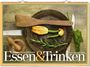"Essen & Trinken" steht groß im Vordergrund. Ein Holzlöffel, Zucchiniblüten und Gewürze auf einer Pfanne arrangiert.