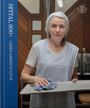 "SPITAL 800+ LEBEN ARBEIT KULTUR". Eine Frau mit Haarnetz und Schürze hält ein Tablett mit einem Tuch in einem Küchenraum.