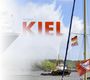 Text: "Peter Schuster", "KIEL", "www.sfk-kiel.de". Ein Schiff auf dem Wasser, Flaggen und Wasserfontänen im Hintergrund.