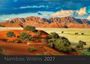 Namibias Wildnis 2027. Rote Sanddünen mit grünen Bäumen im Vordergrund, dahinter felsige Berge und blauer Himmel.