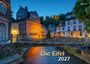 "Die Eifel 2027." Historische Gebäude entlang eines Flusses, beleuchtet im Abendlicht. Ein romantisches Dorfambiente.