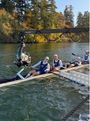 Ruderer in einem Boot auf einem Fluss, umgeben von Bäumen im Herbst. Ein Kamerakran ragt über das Boot.