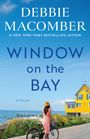 Titel: "Debbie Macomber: Window on the Bay". Frau in Hut neben Rad, blickt aufs Meer. Gelbes Haus, blauer Himmel.